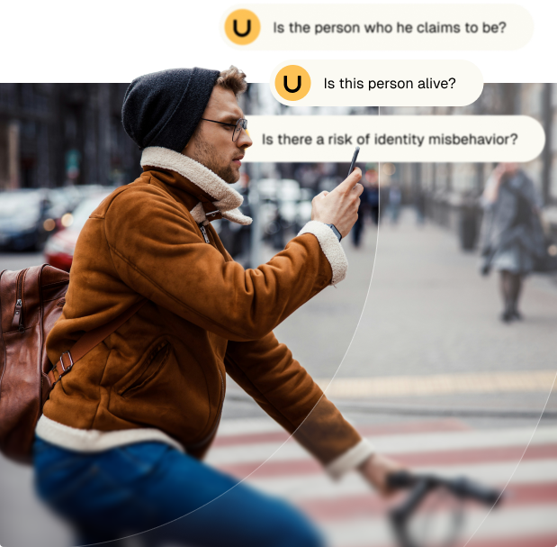Man wearing a brown jacket, glasses, and a beanie rides a bicycle while looking at his smartphone on a city street.