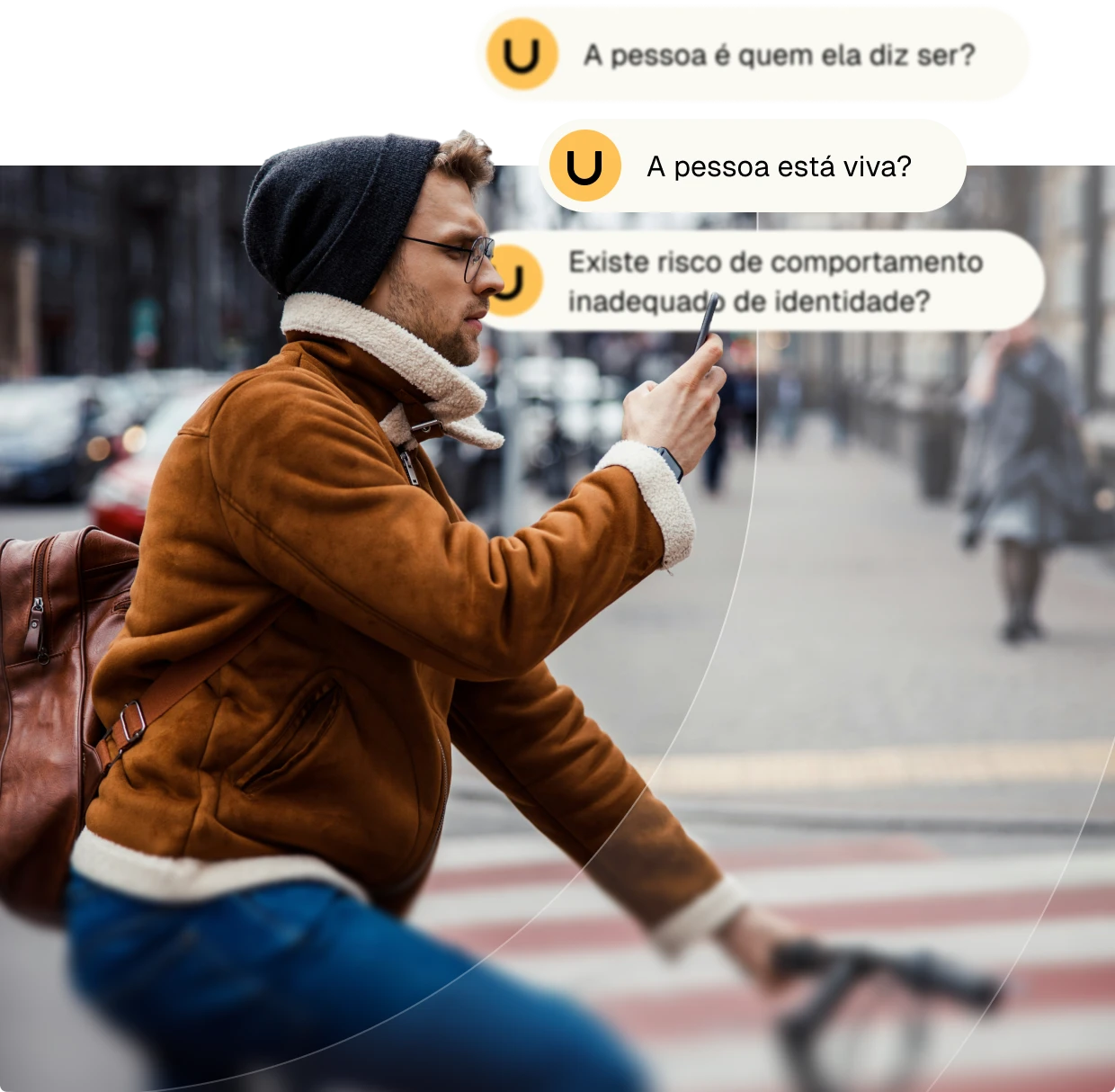 Man wearing glasses, a brown jacket, and a beanie rides a bicycle while looking at his smartphone on a city street.