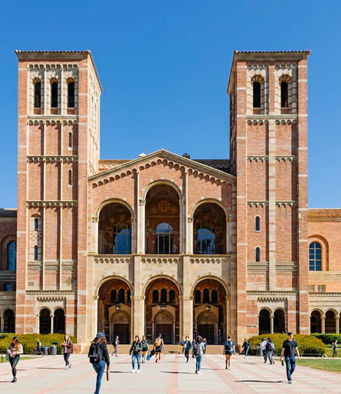 A large, symmetrical brick building with two tall towers and arched windows; people walking in front on a sunny day; the setting suggests a university or academic environment.
