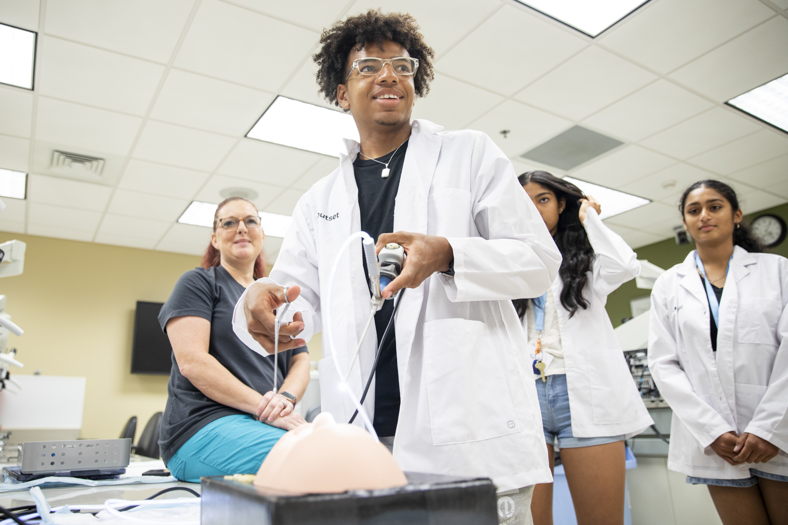 A future medical student in a lab coat manipulates a medical device attached to a silicone model, watched by a seated woman and two standing individuals, in a brightly lit laboratory.