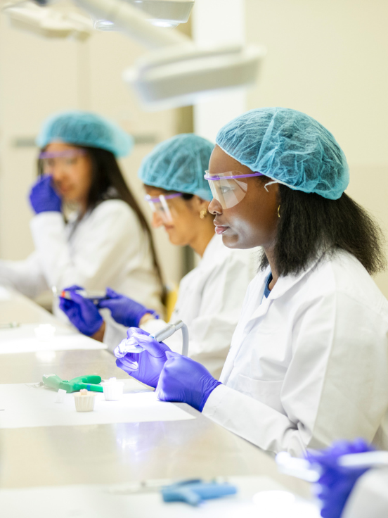Students wearing lab coats and hairnets work at a laboratory bench, focusing on experiments with tools. The environment is well-lit and professional, with three individuals engaged in scientific tasks.