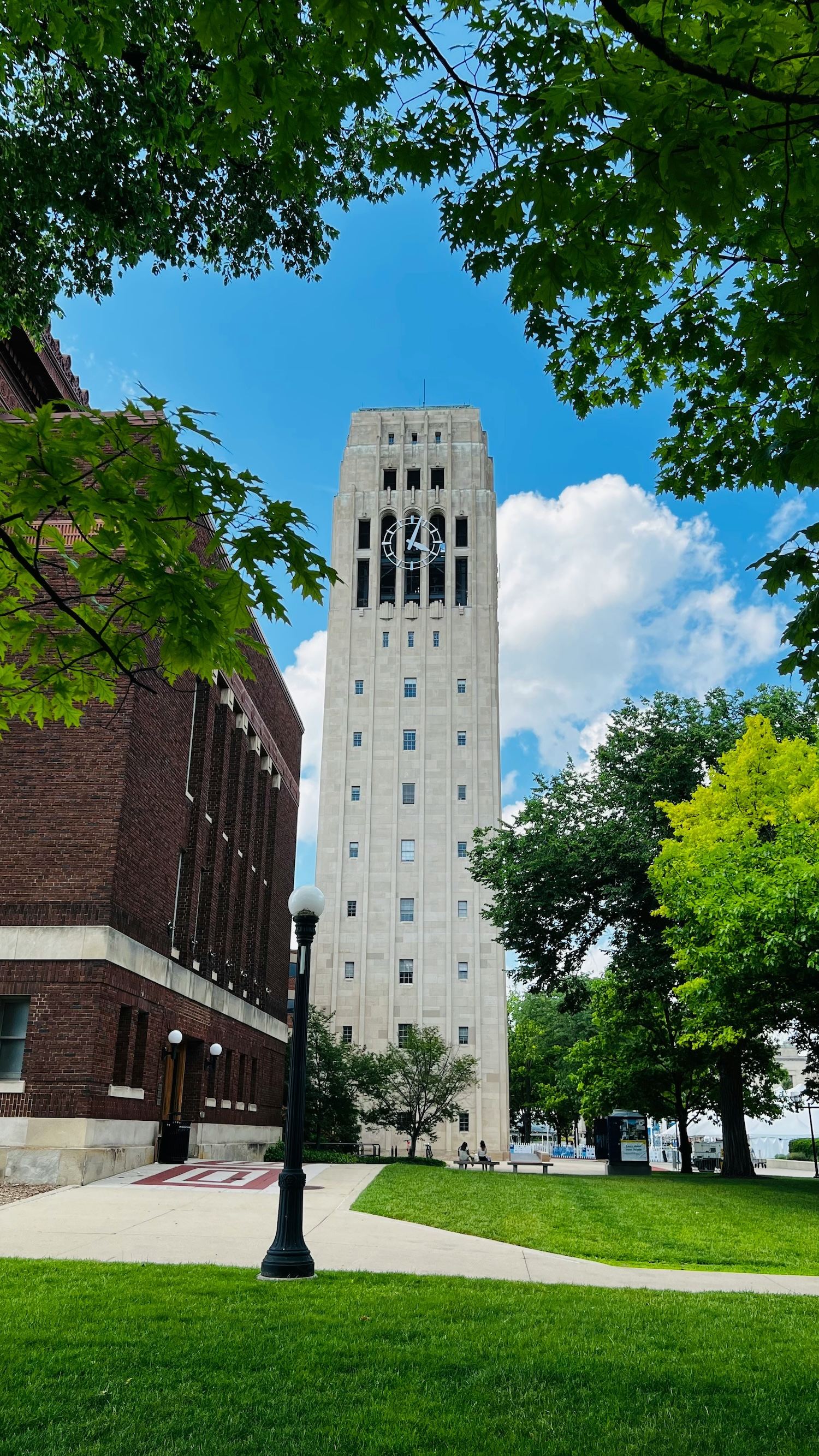 A tall clock tower stands prominently, framed by leafy green trees, next to a brick building under a blue sky with scattered clouds. A lamppost and green lawn complete the scene.