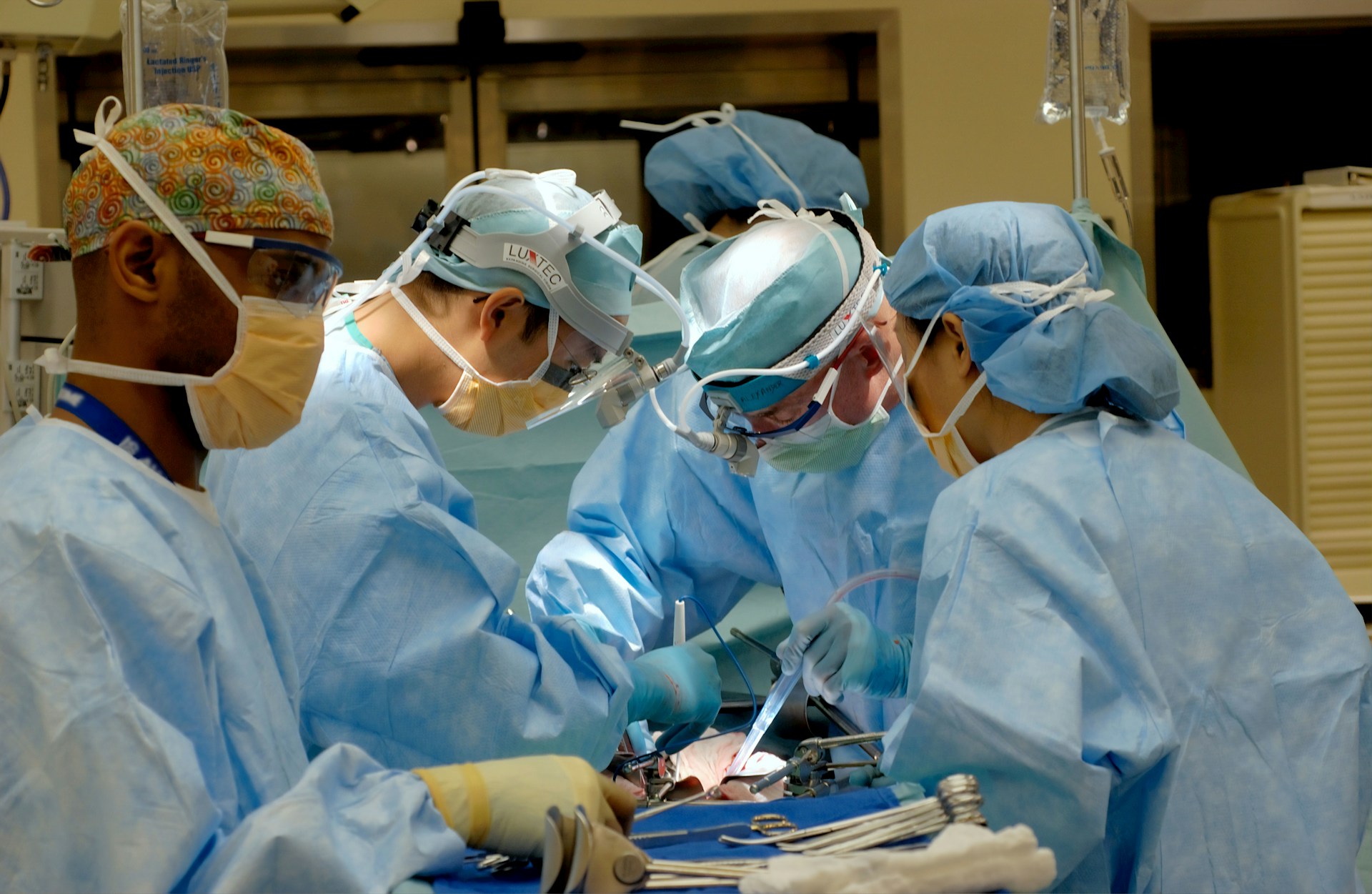 Surgeons perform a medical procedure, focused intently on a patient. They wear blue surgical gowns, masks, and caps in a sterile operating room, surrounded by medical instruments.