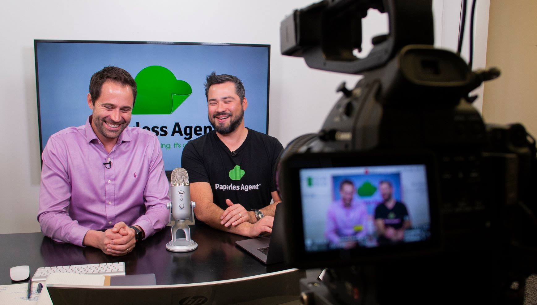 Two men sitting at a desk with a microphone and laptop, filming a video with a camera in front; a screen behind shows the Paperless Agent logo.