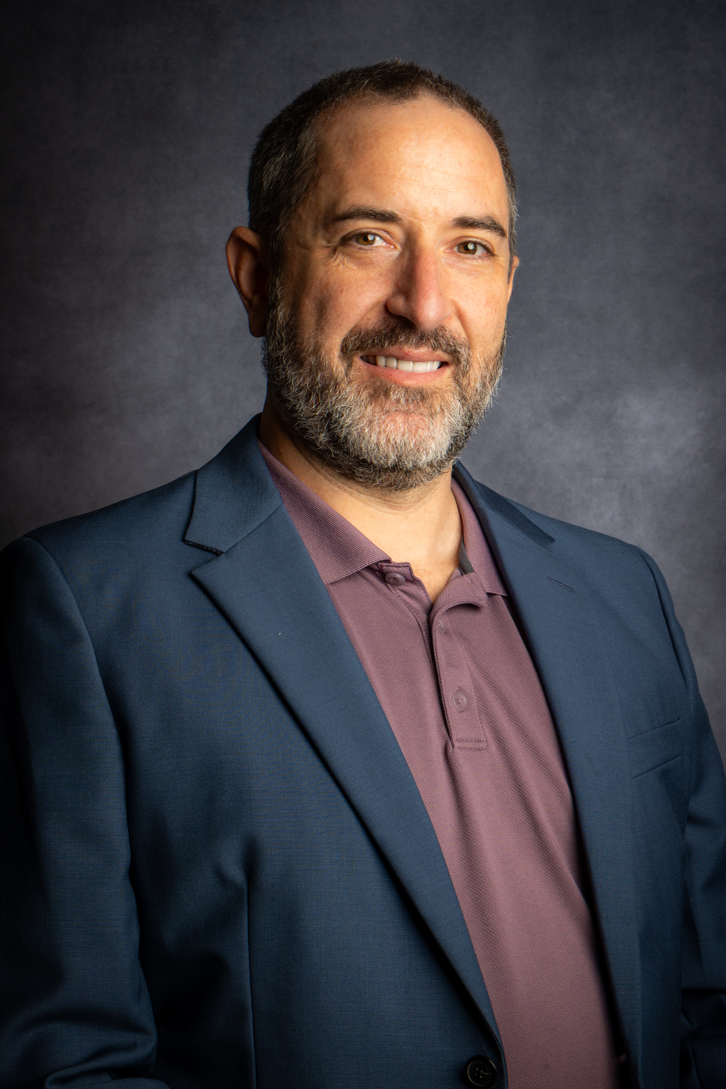 Middle-aged man with salt-and-pepper beard wearing a blue blazer over a maroon polo shirt, smiling against a dark gray background.