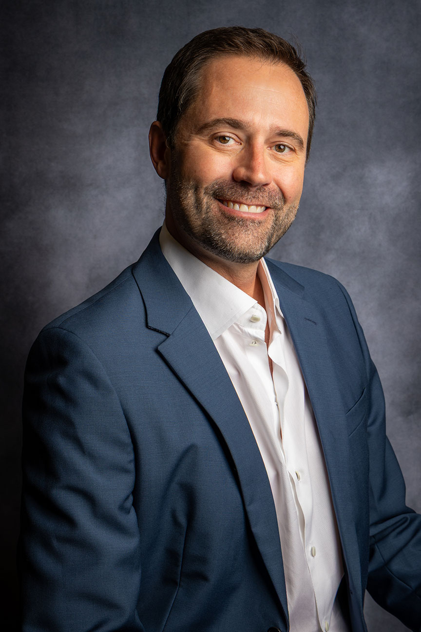 Smiling man with short dark hair and beard wearing a blue blazer and white shirt against a dark gray background.