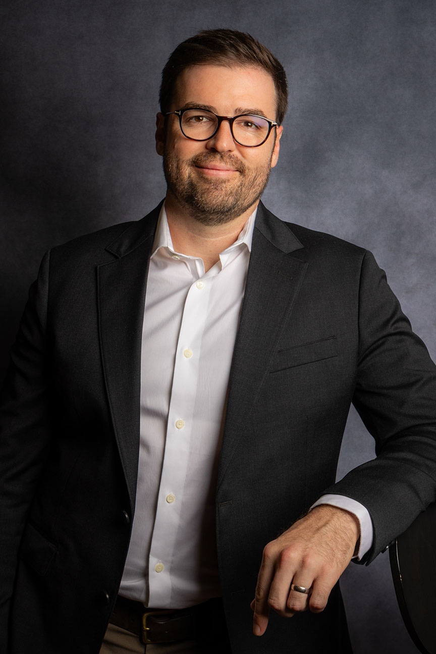 Smiling man with beard and glasses wearing a dark blazer and white shirt, posing against a gray background.