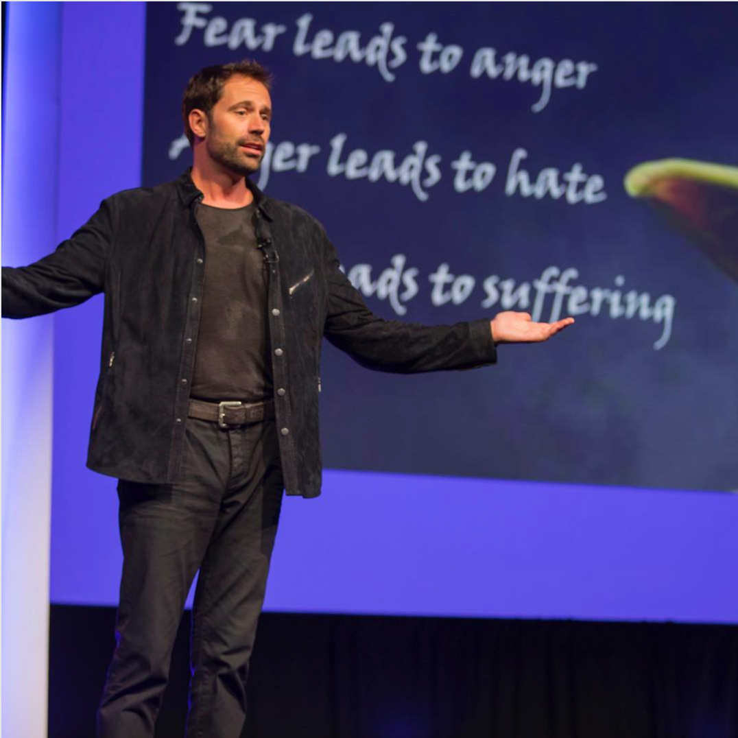 Man in black jacket and shirt speaking on stage with text on screen behind reading 'Fear leads to anger, anger leads to hate, leads to suffering.'