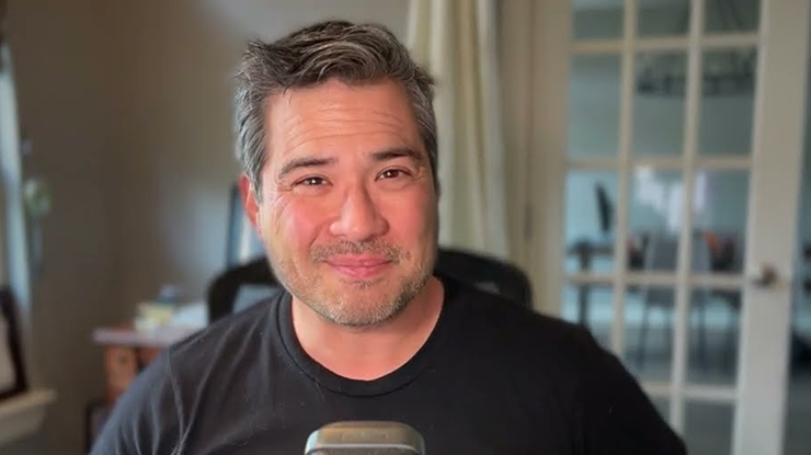 Smiling man with salt-and-pepper hair sitting indoors in front of a microphone and blurred office background.