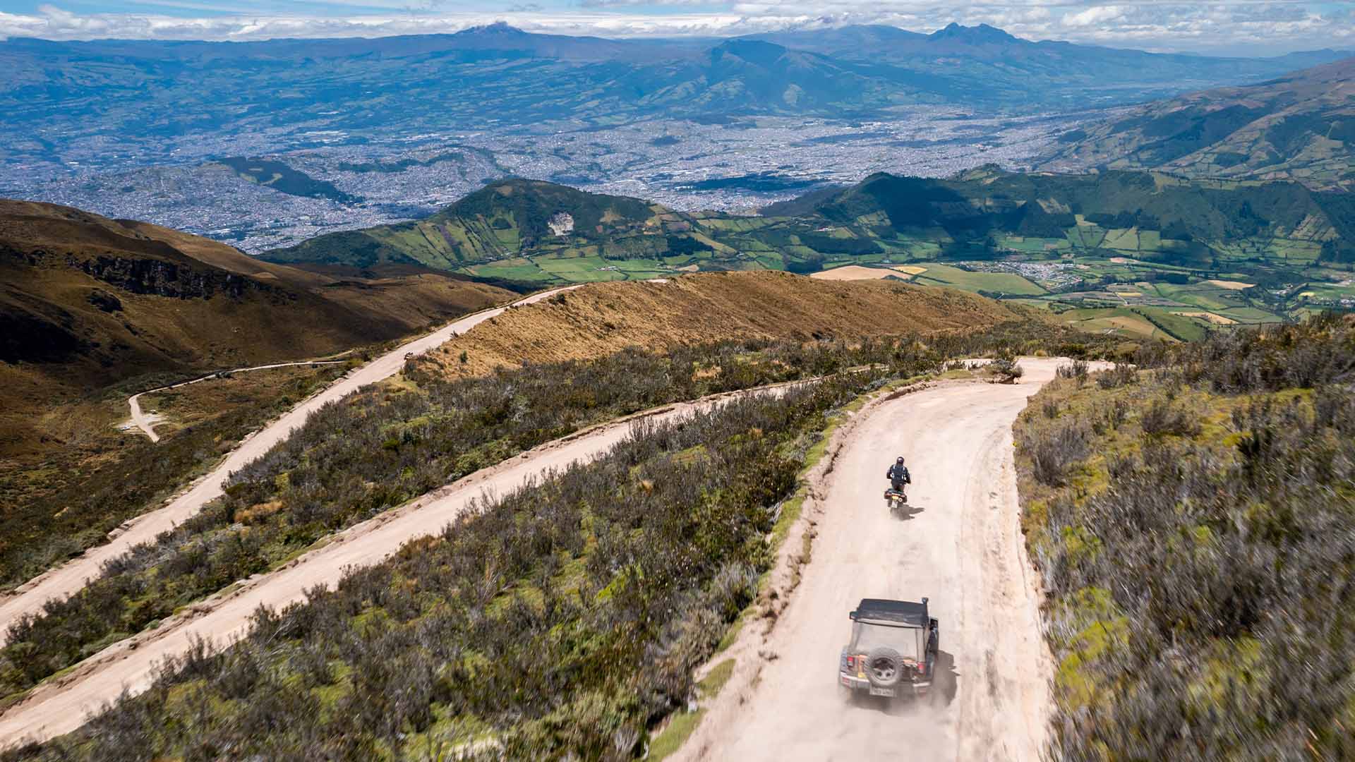 A set of dirt roads converging in a mountainous terrain from an aerial view with a motorcyclist and a film crew following behind.