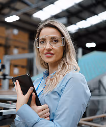 MAU team member in safety glasses holding a phone on an industrial manufacturing floor