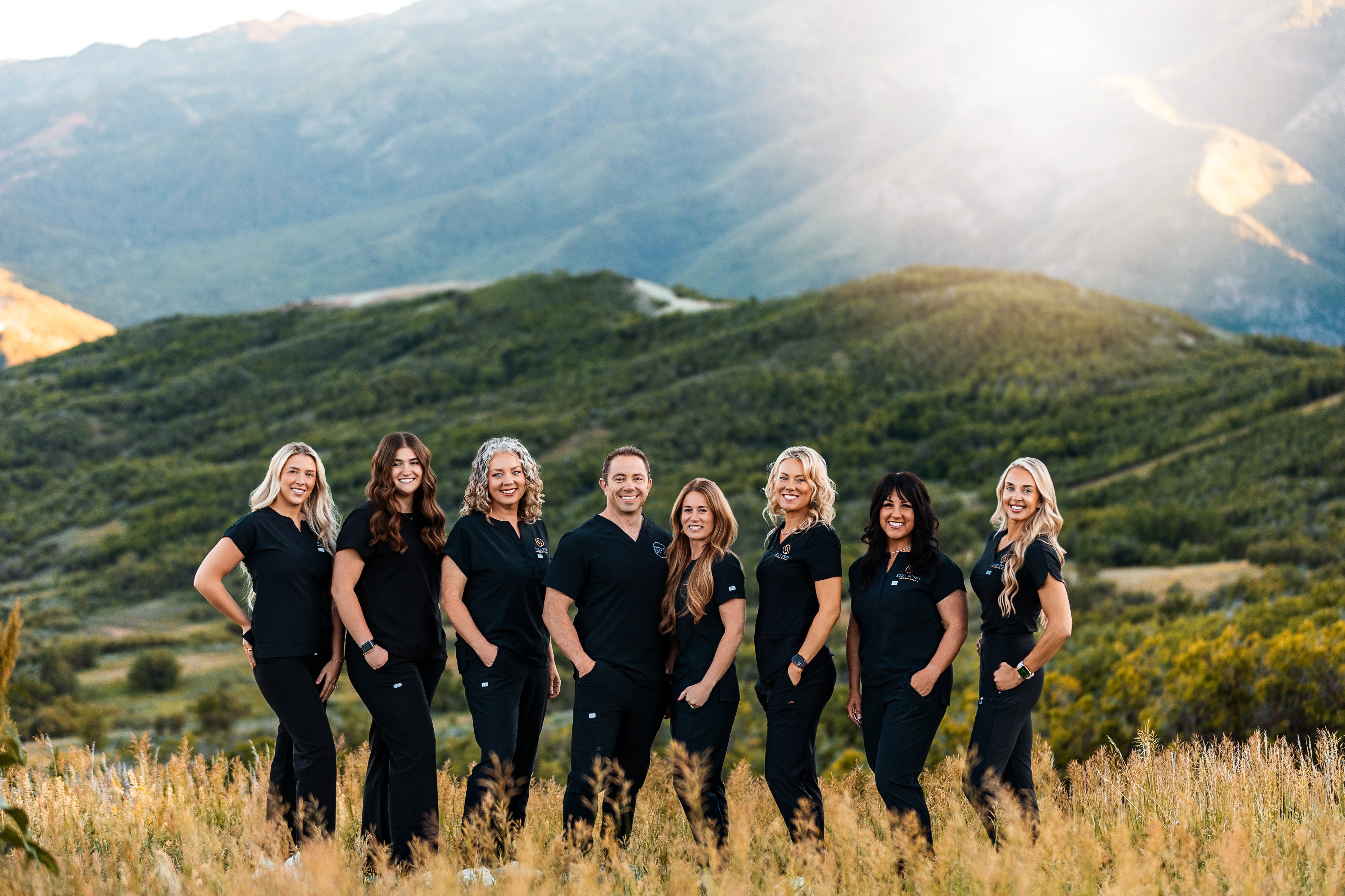 Eight people in black Still Point Medical Spa scrubs standing side by side in a field with mountains behind them.