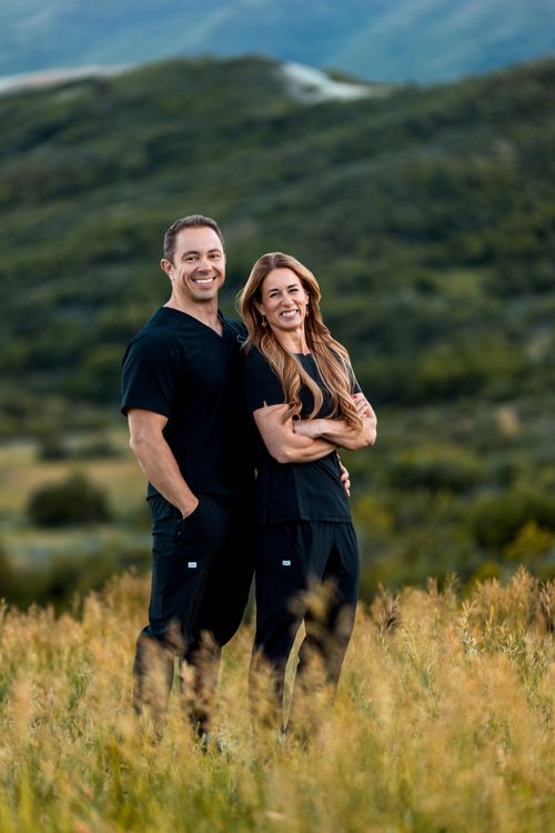 Smiling man and woman wearing black Still Point Medical Spa scrubs standing in tall grass with hills in background.