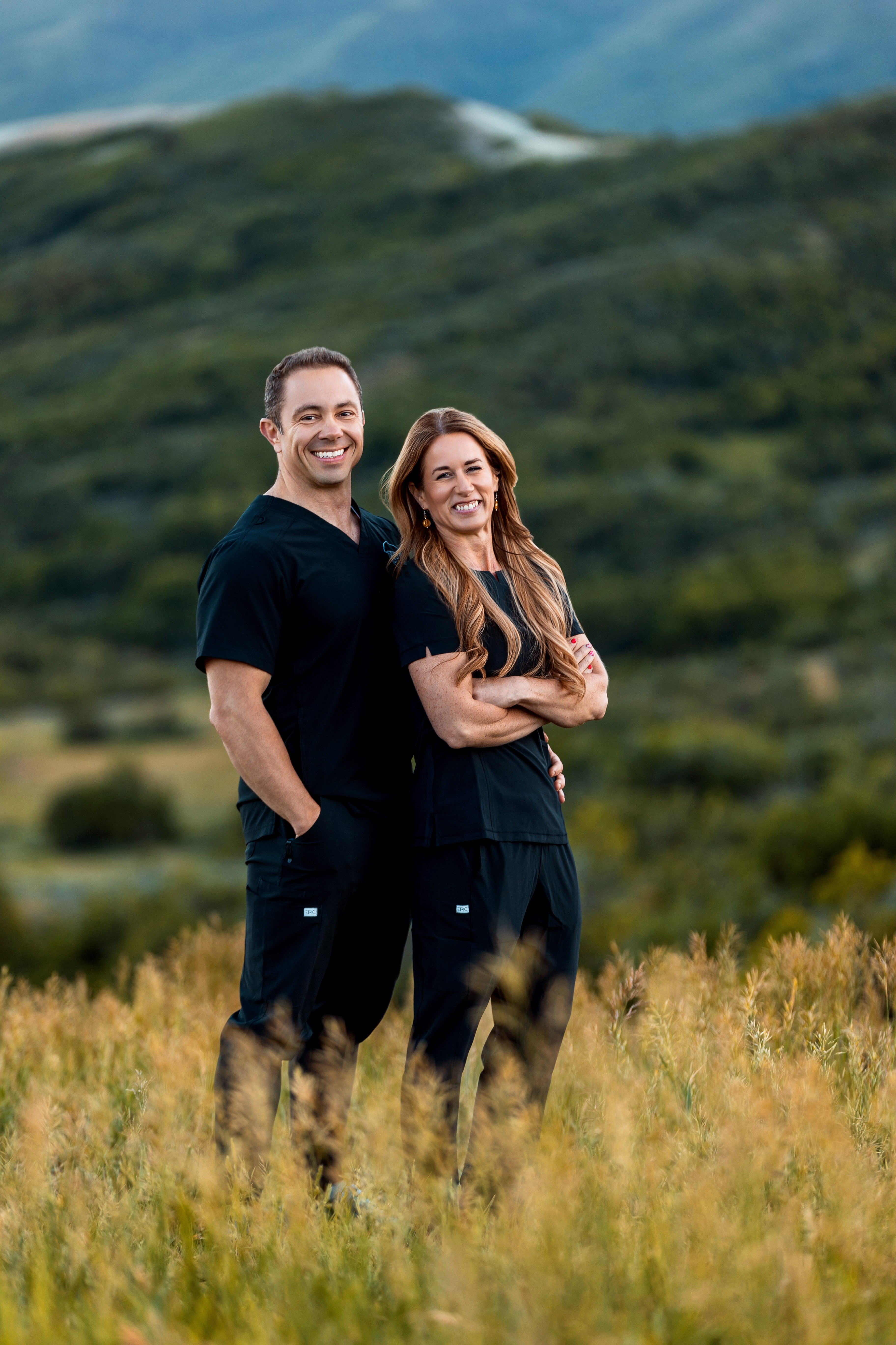 Smiling man and woman wearing black Still Point Medical Spa scrubs standing in tall grass with hills in background.