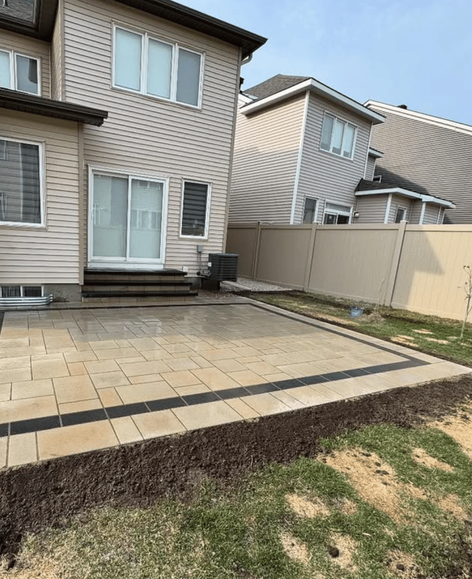 Backyard patio with beige and dark brown rectangular tiles adjacent to a beige house, surrounded by a beige privacy fence.
