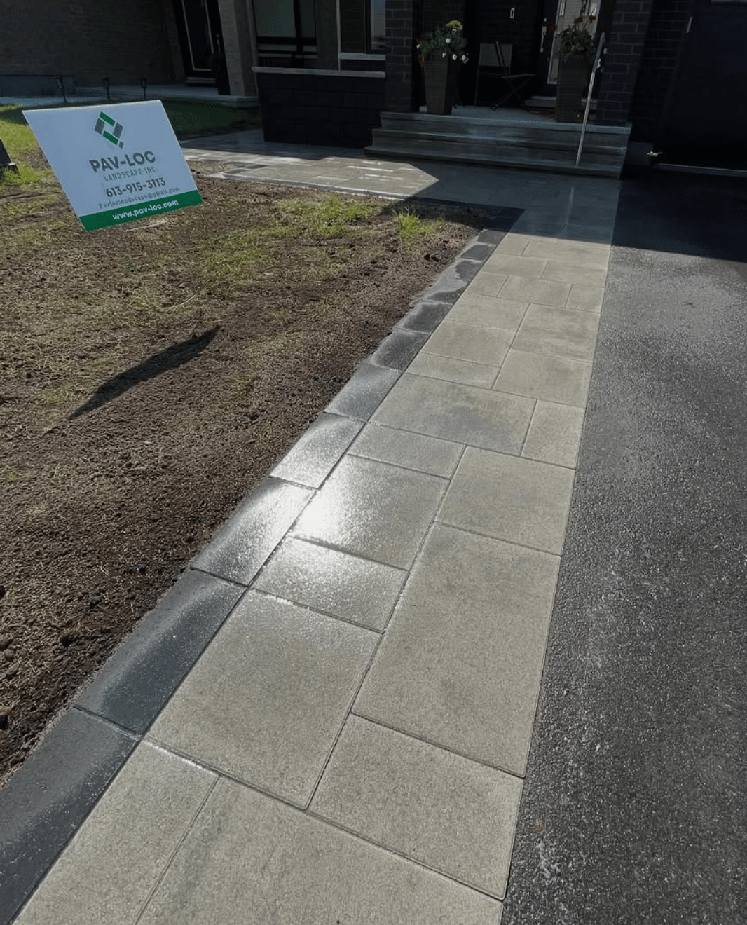 Newly installed beige and gray paving stones forming a walkway beside a driveway and soil area with a Pav-Loc Landscape Inc sign.