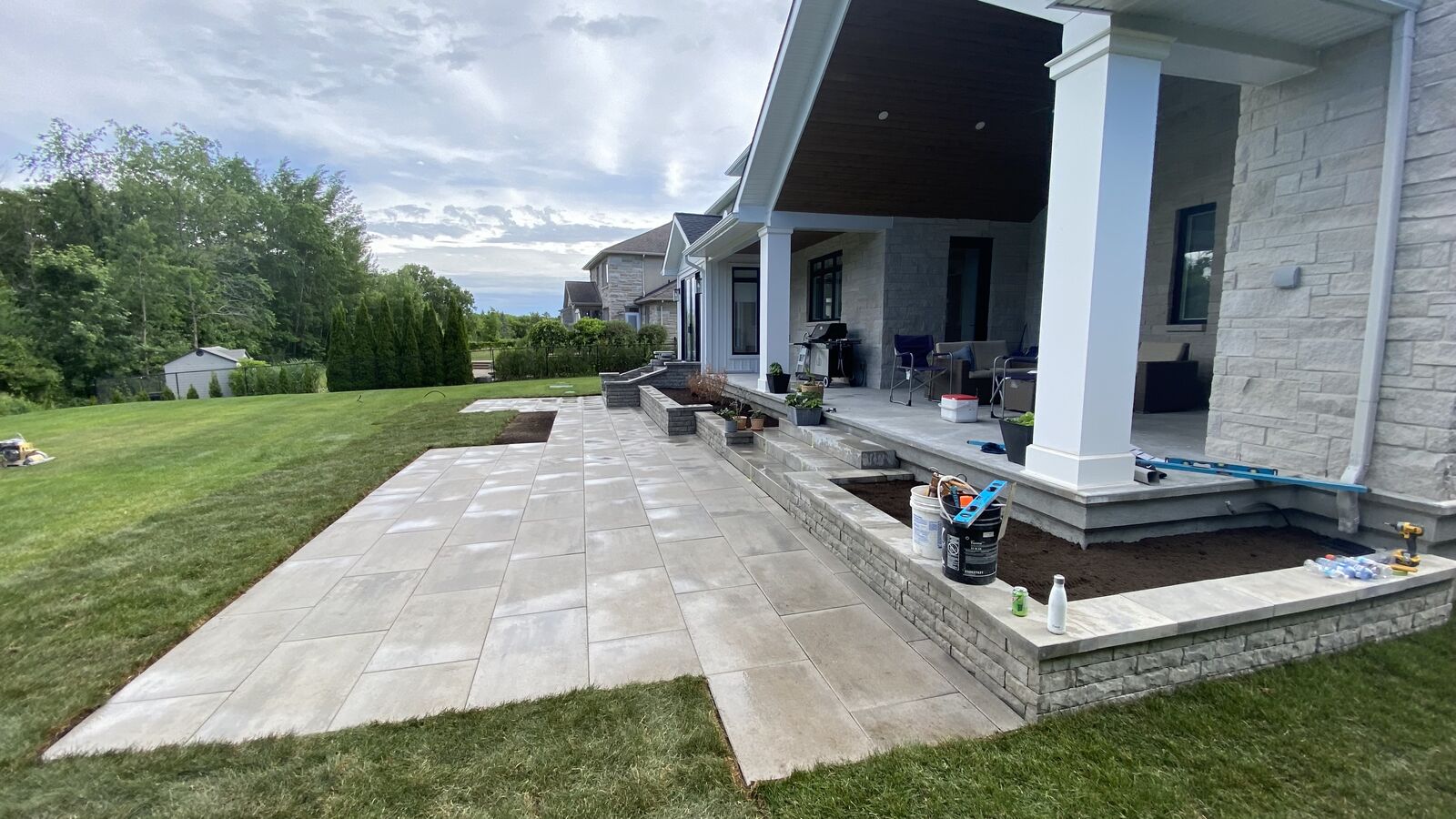 Newly constructed outdoor patio area with large light gray tiles, raised stone flower beds, and a covered porch attached to a light stone house with outdoor furniture and gardening supplies.