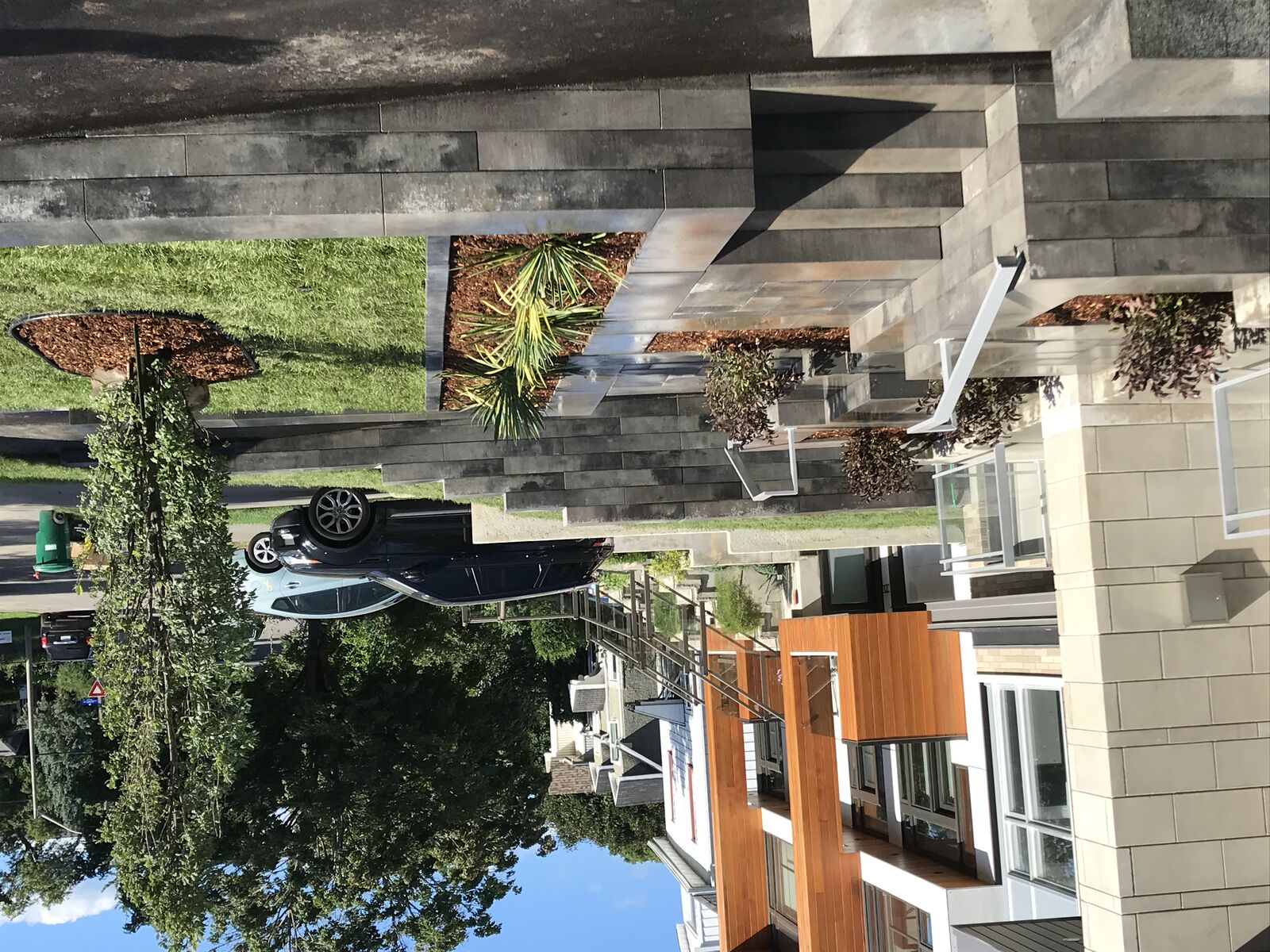 Modern residential front yard with stone steps, raised flower beds, green grass, and small trees and shrubs.