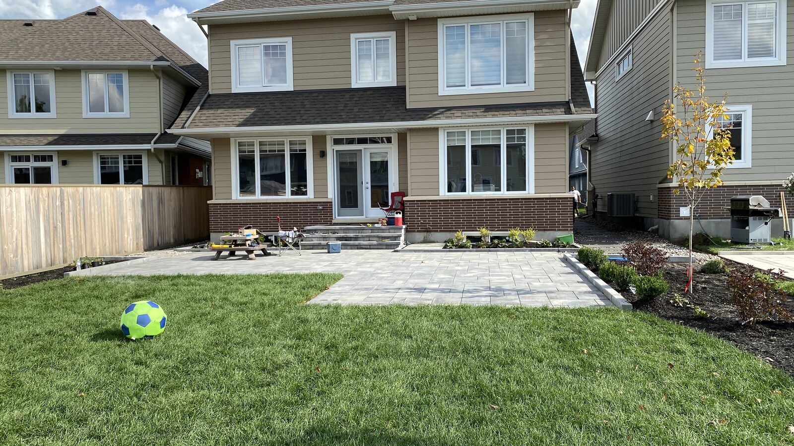 Backyard of a suburban house with a green lawn, a blue and green soccer ball, a paved patio area, a small tree, and a wooden fence.