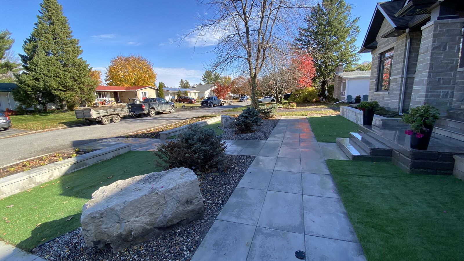 Modern sidewalk and front yard landscaping with stone steps, green grass patches, trees, shrubs, and a large rock under a clear blue sky.