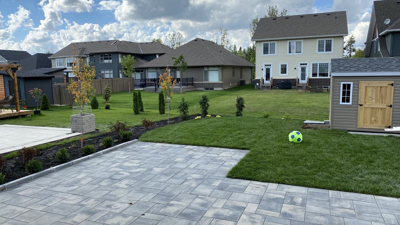 Backyard with a paved patio, green lawn, small trees, shrubs, a soccer ball, and a wooden shed under a partly cloudy sky.