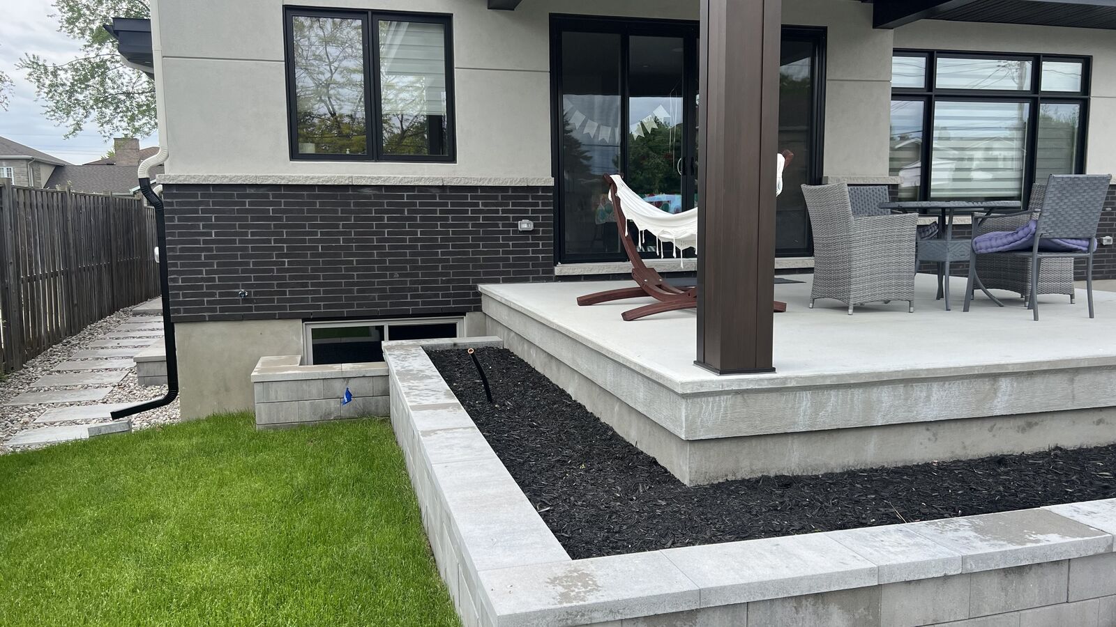 Backyard patio with a concrete raised platform supporting a hammock and outdoor wicker chairs around a table, adjacent to a grass lawn and a raised garden bed filled with mulch.