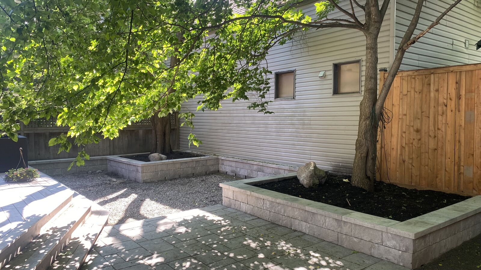 Backyard patio with stone steps, paved surface, raised garden beds with trees, and a wooden fence.