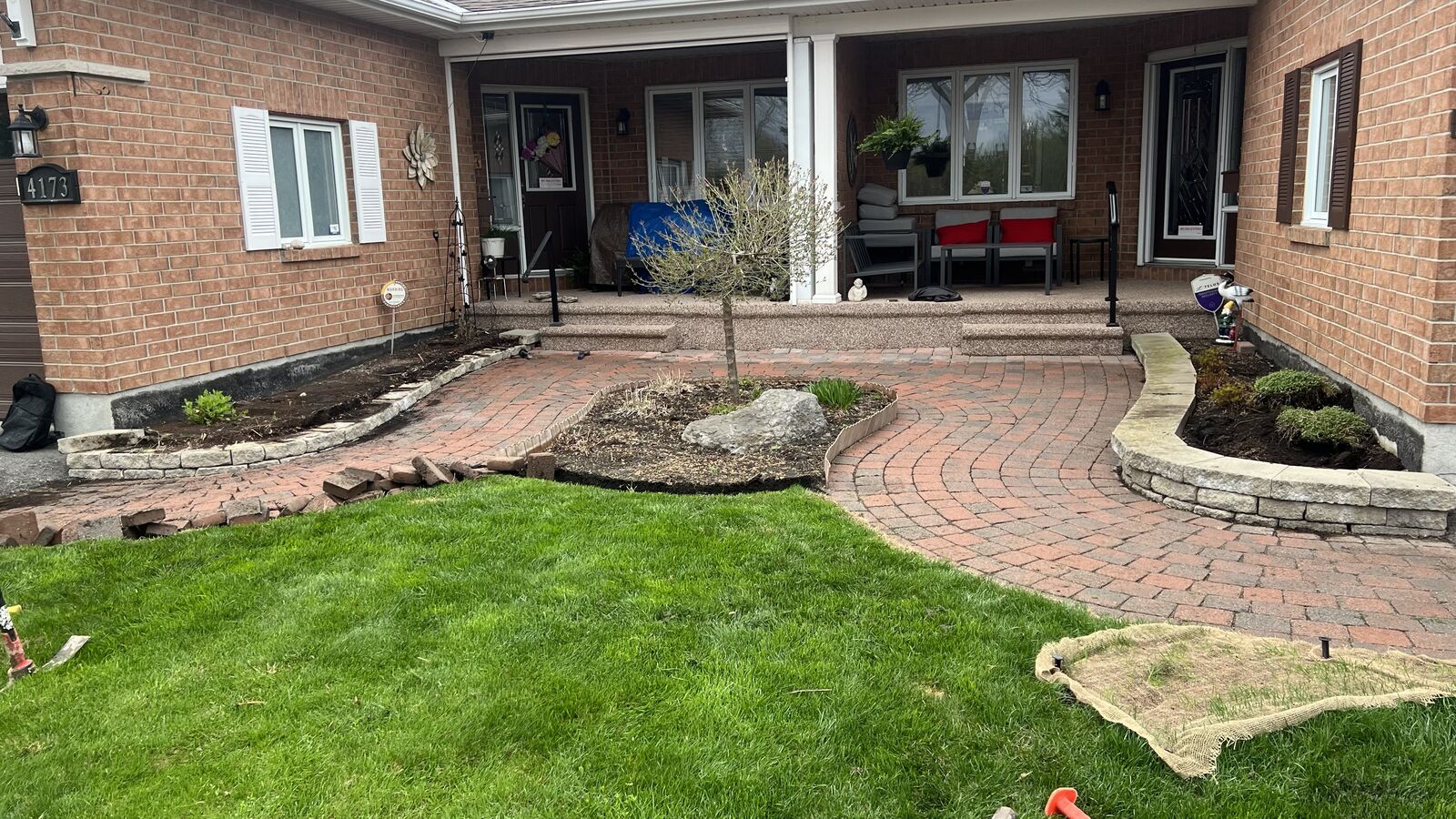 Front yard of a brick house with a curved brick walkway, a small leafless tree in a mulched bed, and garden beds along the walls.