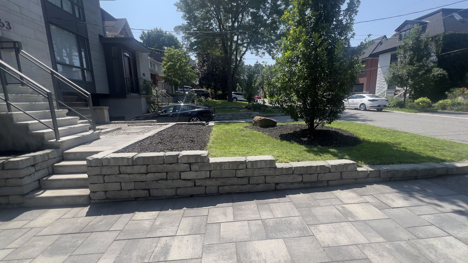 Modern front yard with stone retaining wall, paved walkway, grassy lawn, tree, and steps leading to house entrance.