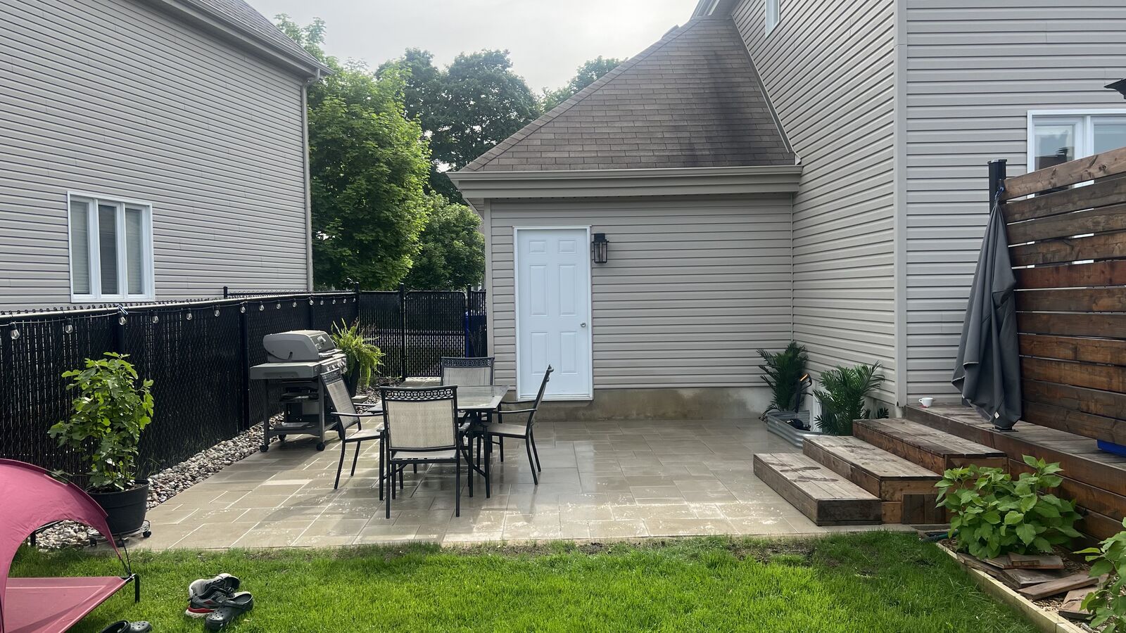 Backyard patio area with tiled flooring, outdoor dining table with four chairs, grill, wooden steps, and surrounding green plants and grass.