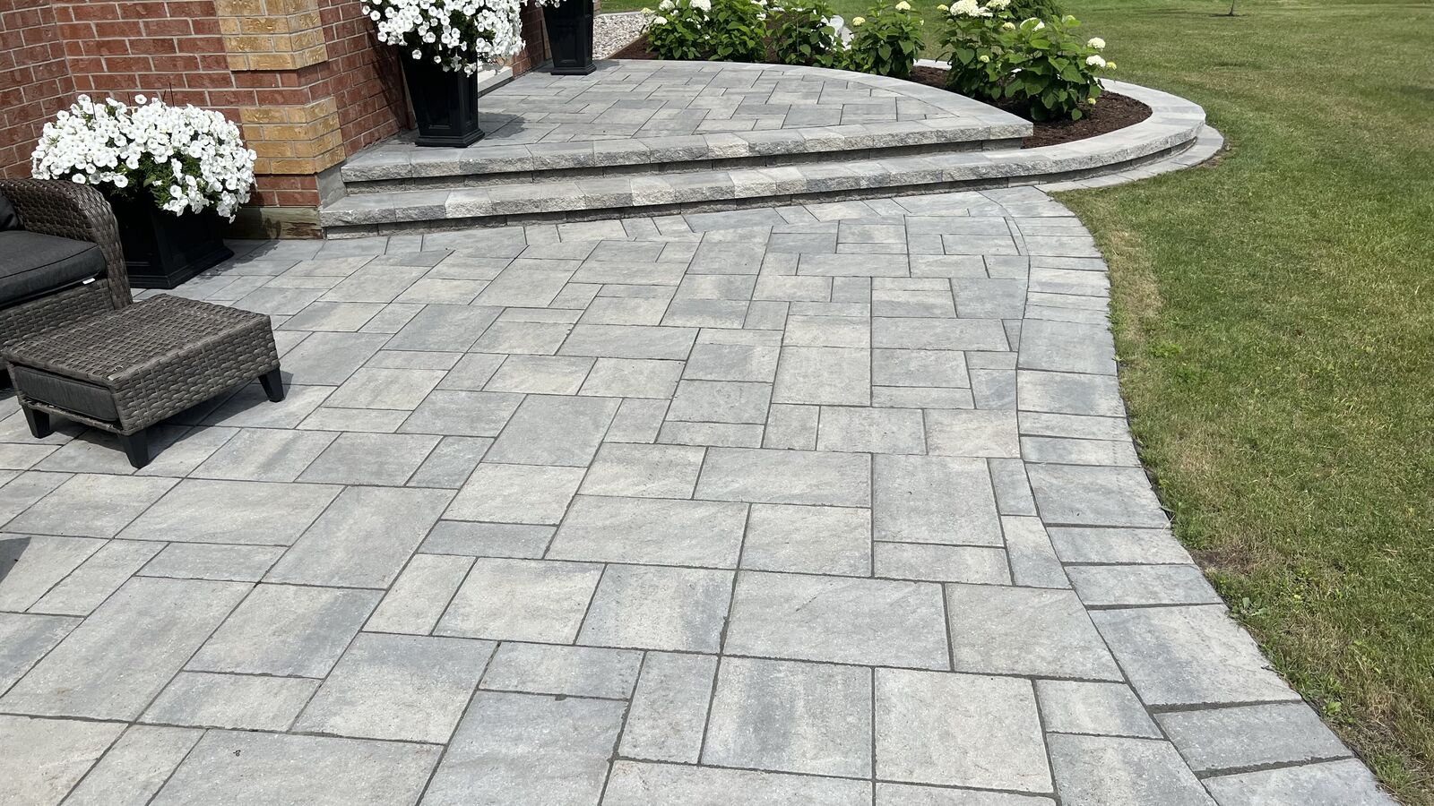 Outdoor stone patio with a curved stairway, potted white flowers, wicker chair, and adjacent green lawn.