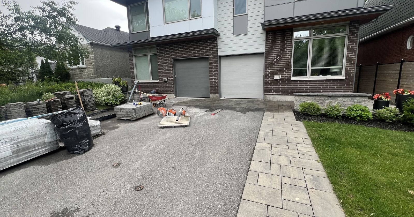 Driveway under construction with stacked paving stones, wheelbarrow, and tools in front of a modern brick house with two garage doors.