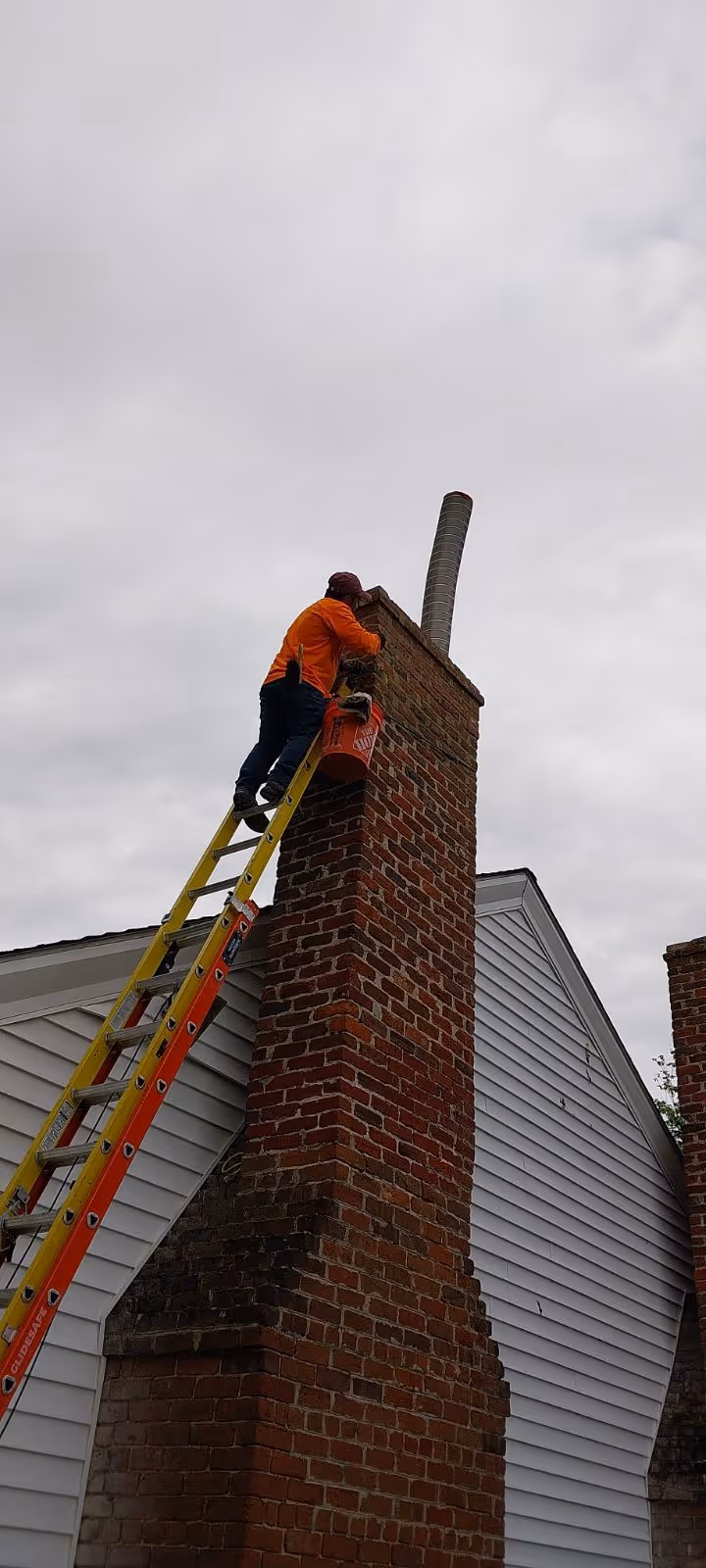 Worker in an orange jacket on a yellow ladder repairing a brick chimney on a house with gray siding under a cloudy sky.