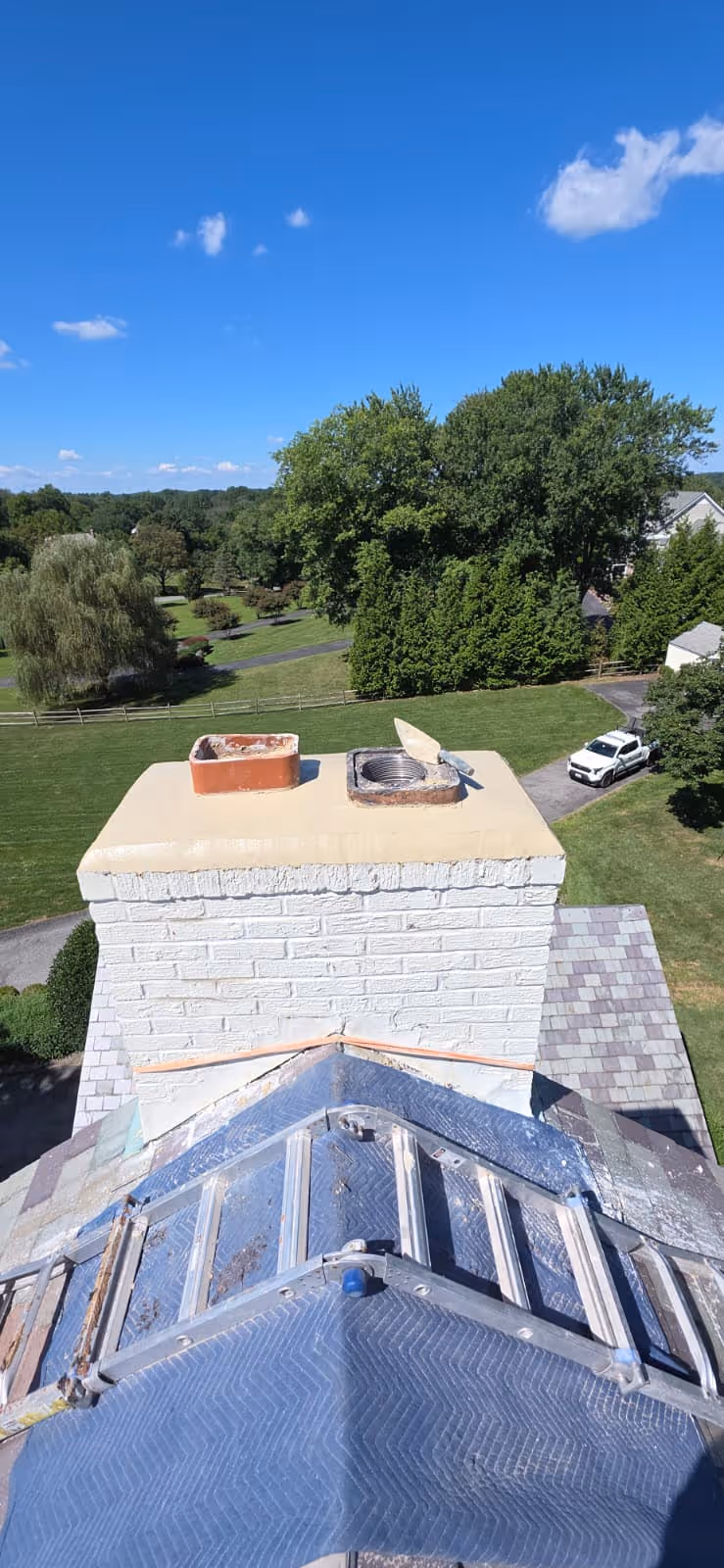 View from rooftop showing a white brick chimney with two flue openings and green trees and a white pickup truck in the background under a clear blue sky.