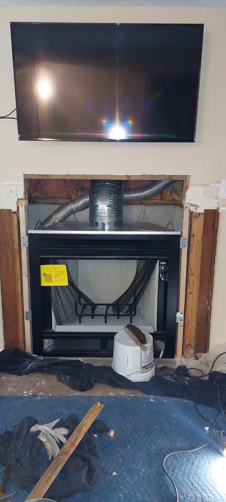A modern black fireplace insert installed in a wall opening under a wall-mounted TV, with exposed ductwork above and tools scattered on a blue protective floor covering.