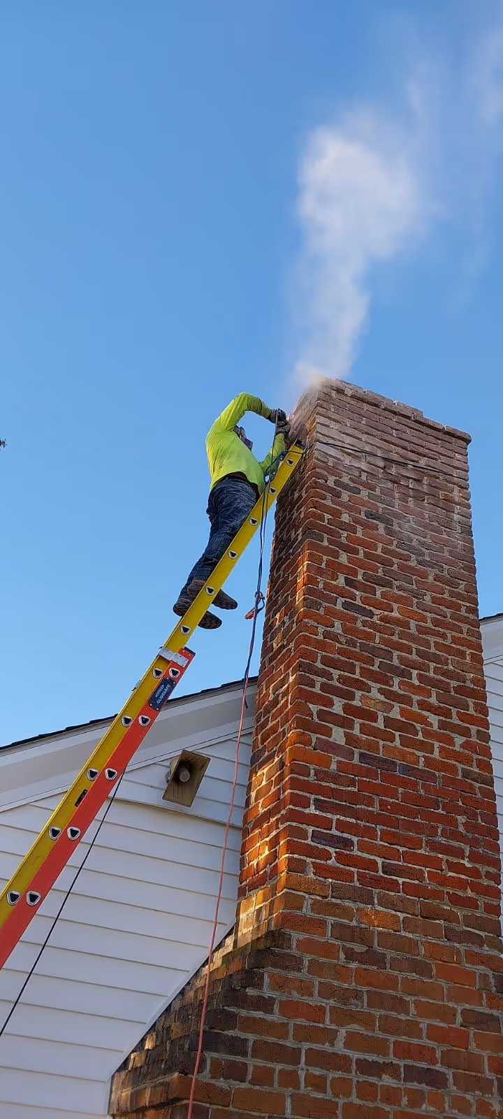 Worker in a bright green jacket standing on a yellow ladder cleaning or repairing a brick chimney with smoke rising against a clear blue sky.