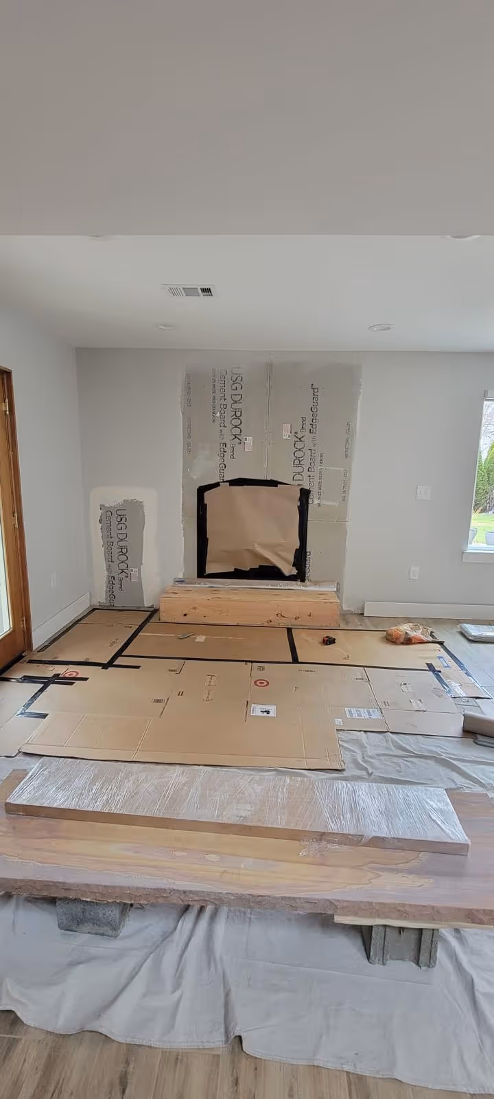 Interior room under renovation with protective cardboard on the floor and cement boards on the wall surrounding a fireplace opening covered with brown paper.