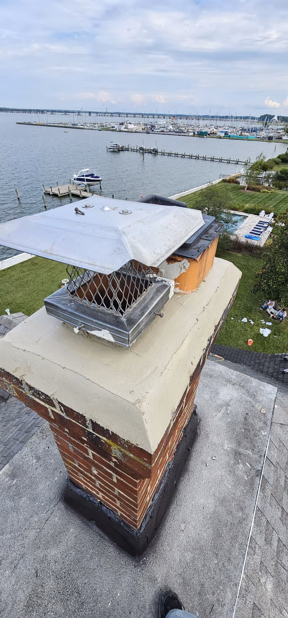 Brick chimney with metal chimney cap on a rooftop overlooking a waterfront with boats, docks, and a lawn with a swimming pool.