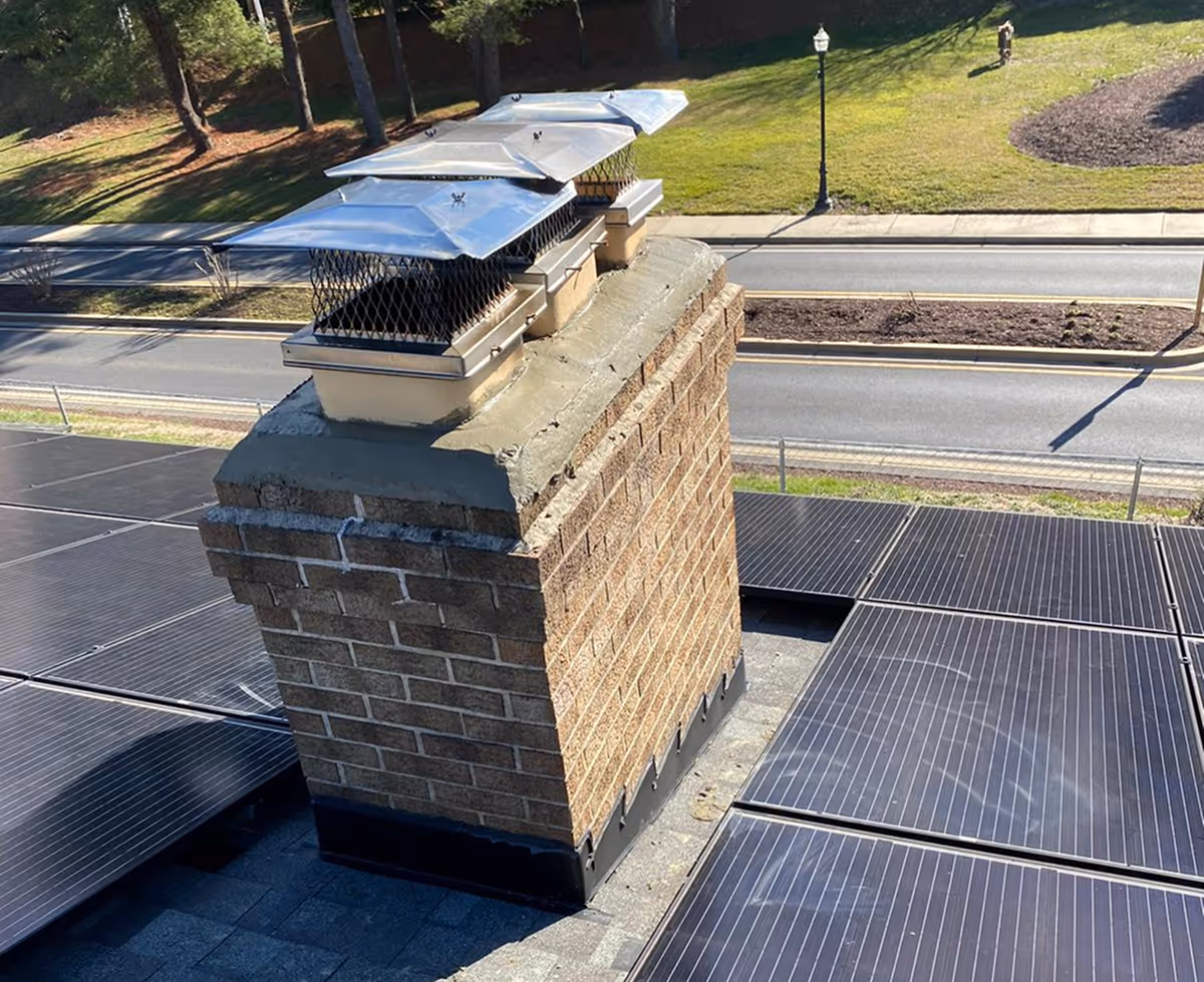 Brick chimney with metal caps on a rooftop surrounded by solar panels, a road, and grassy area with trees in the background.