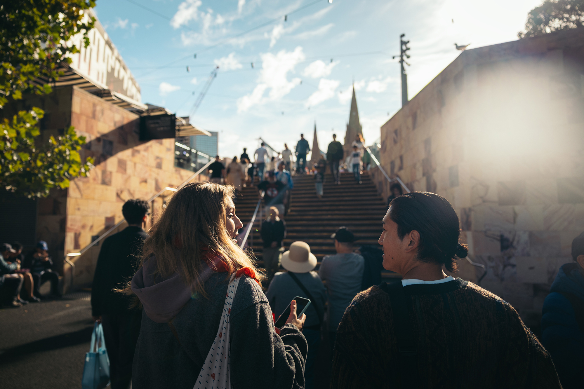 Two people sitting across from each other at a table working on laptops with a large green plant and window in the background.