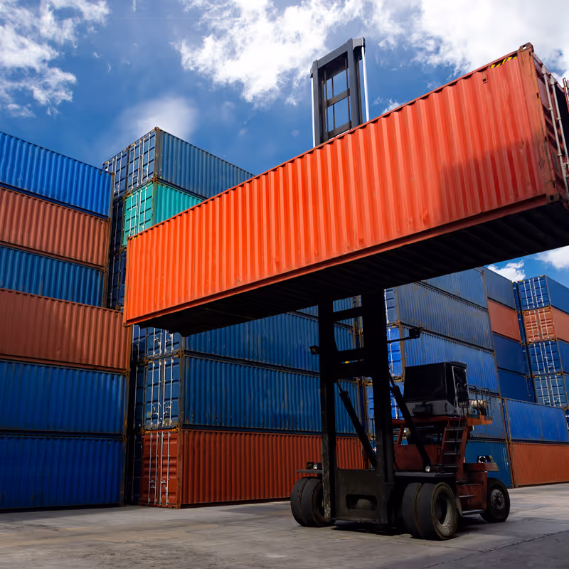 Forklift lifting a large red shipping container in a yard filled with stacked blue and red containers under a partly cloudy sky.