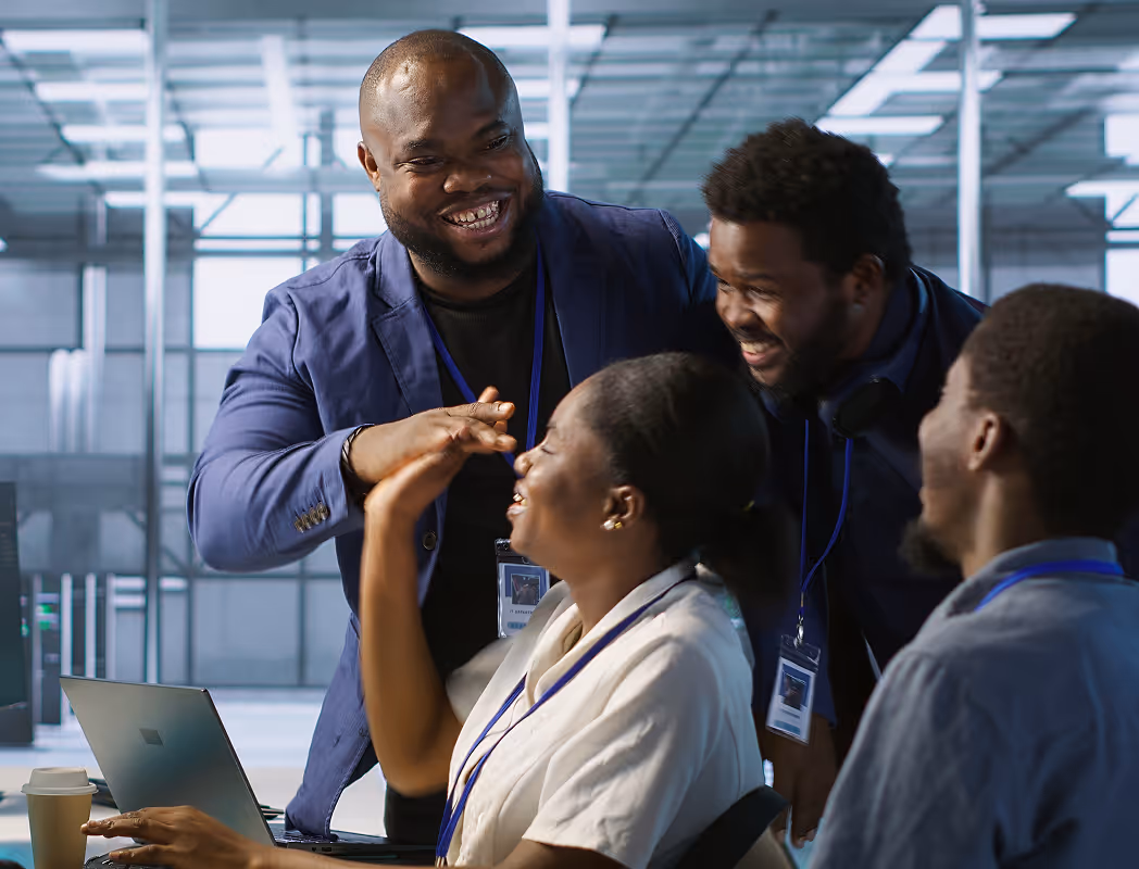 Four colleagues in an office laughing and giving a high-five around a laptop.