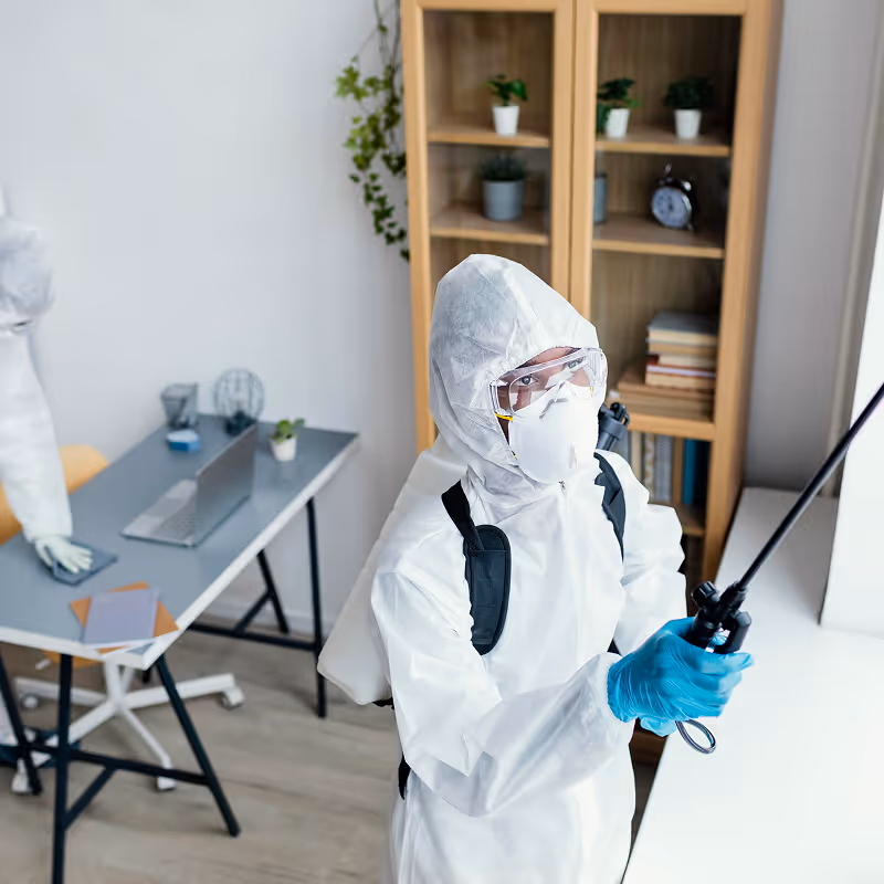 Person wearing protective suit, mask, goggles, and gloves spraying disinfectant in a room with a desk and bookshelf.