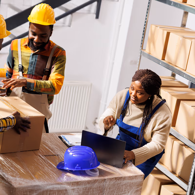 Man in a yellow hard hat and colorful shirt handling boxes while a woman in overalls uses a laptop in a warehouse with stacked boxes on shelves.