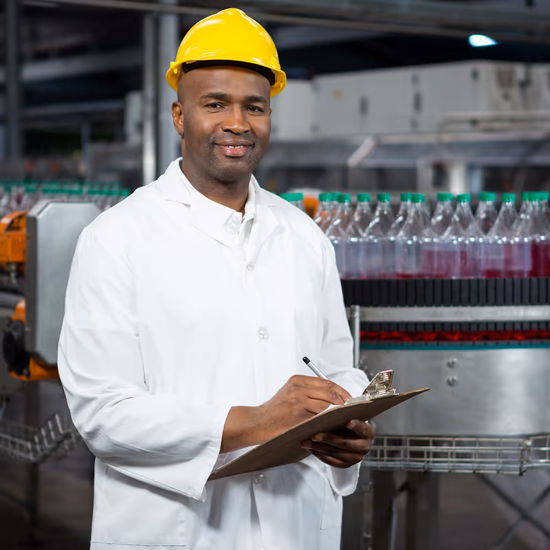 Smiling man in a white lab coat and yellow hard hat holding a clipboard in a beverage bottling factory.