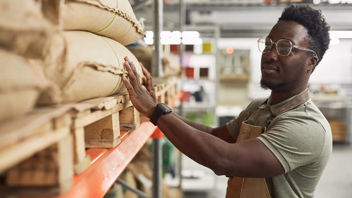 Man wearing glasses and apron inspecting large burlap sacks stacked on warehouse shelves.