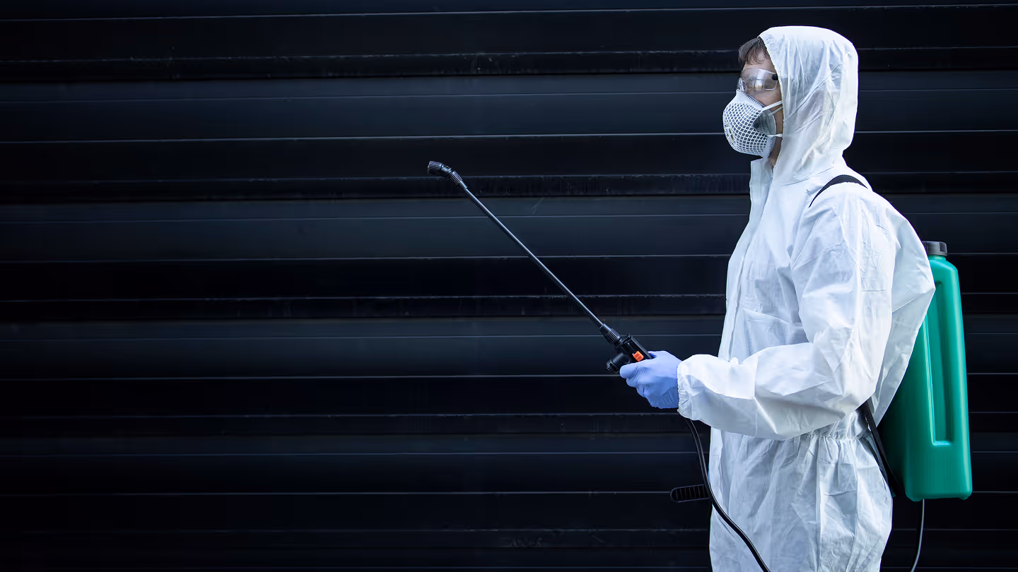 Person wearing white protective suit, gloves, mask, and goggles holding a pest control spray wand with a green tank on their back against a dark background.