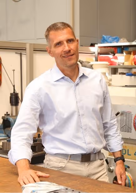 Mann mit Bart und blonden Haaren in blauem Anzug mit verschränkten Armen vor einem modernen Bürofenster.