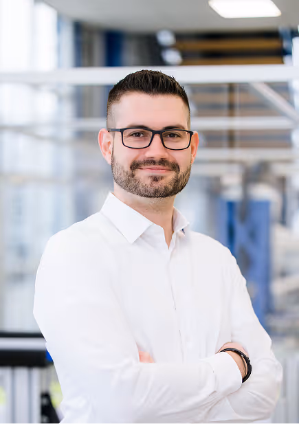 Mann mit Bart und blonden Haaren in blauem Anzug mit verschränkten Armen vor einem modernen Bürofenster.