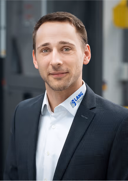 Mann mit Bart und blonden Haaren in blauem Anzug mit verschränkten Armen vor einem modernen Bürofenster.